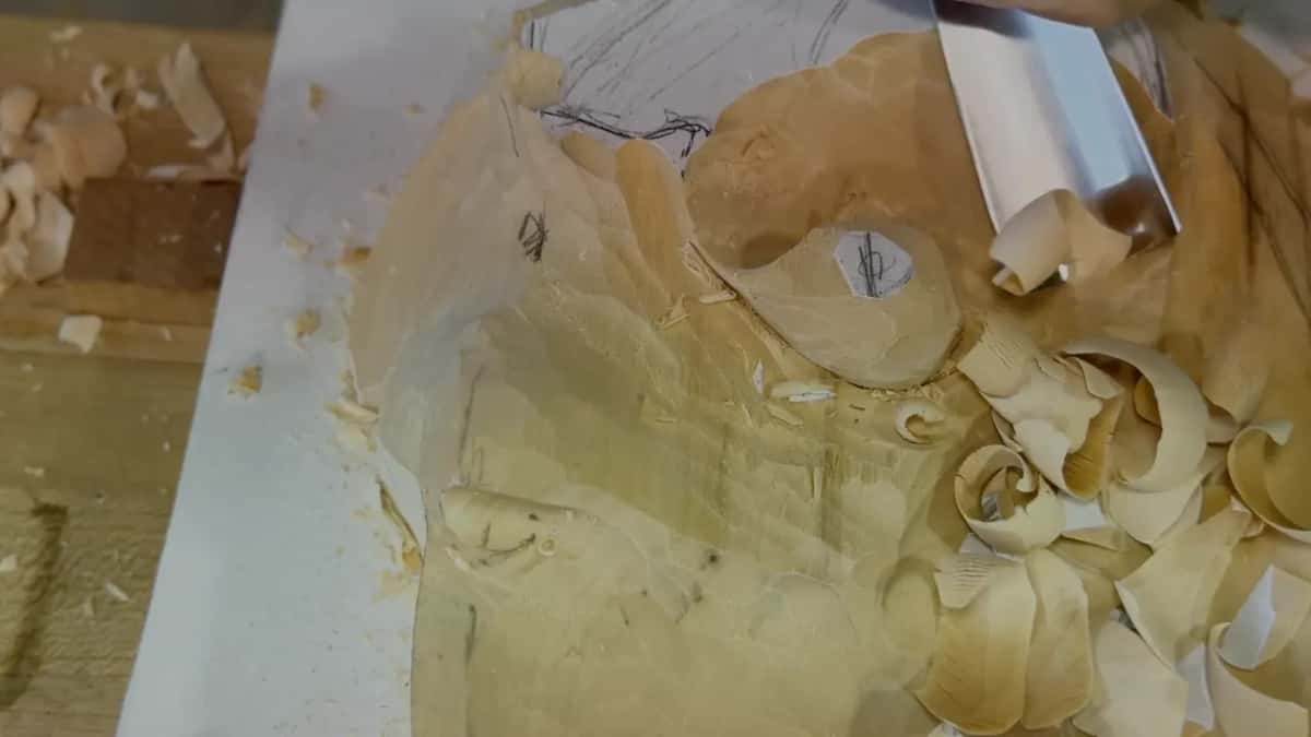 Curled shavings heaped round the half carved leaf, a wide blade at the top, pencil lines on the paper below