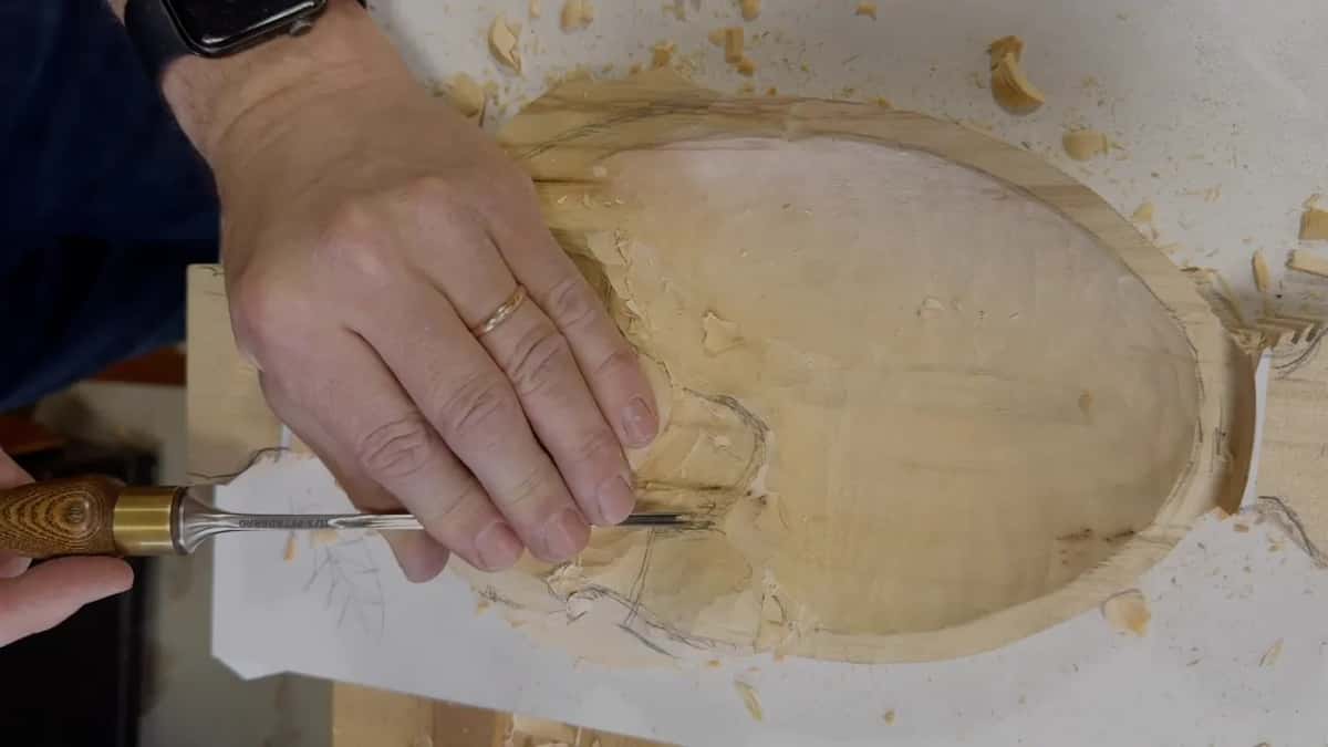 Both hands guiding a slim gouge into the side of the leaf, chips on the paper