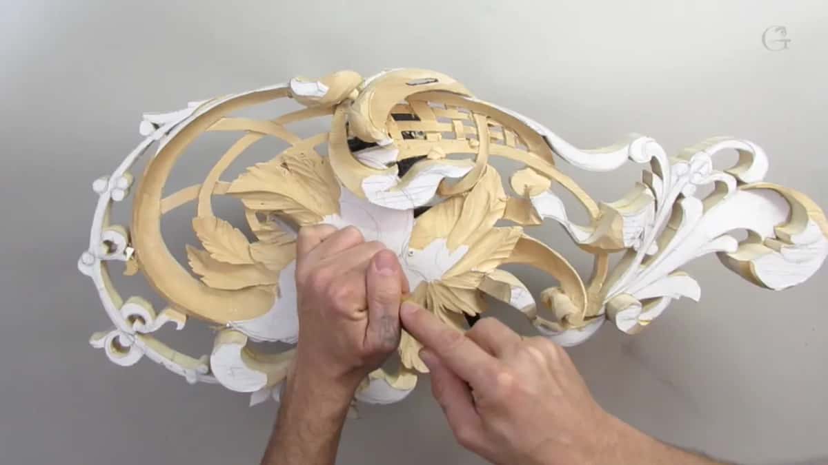 Two hands rest on the oval carving from above, the outer scrolls white and the leaves bare wood.