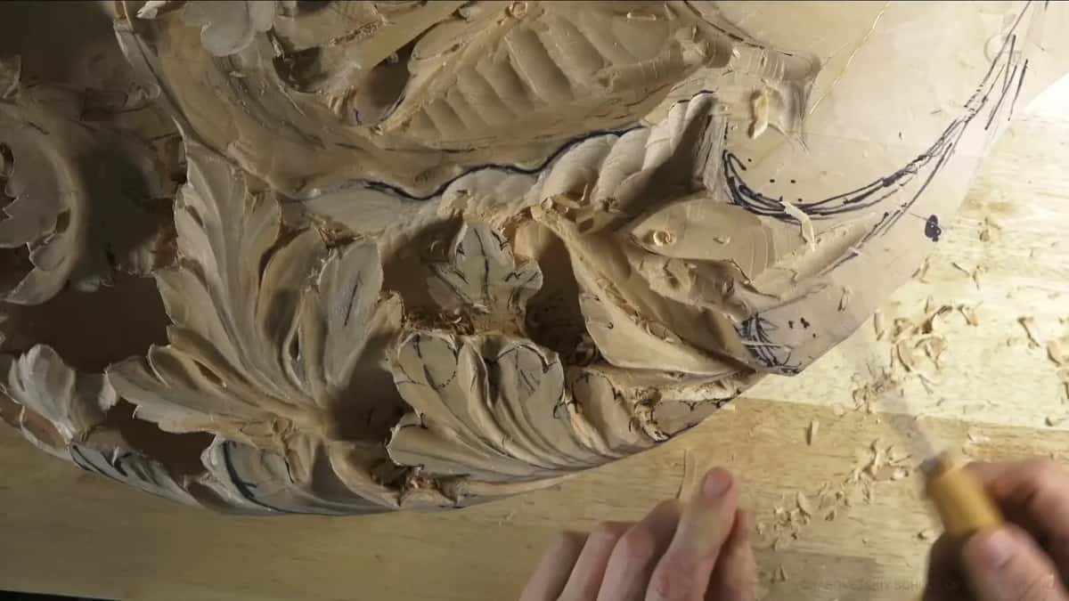Fingers pinch a small pale handled tool right at the edge of a carved leaf