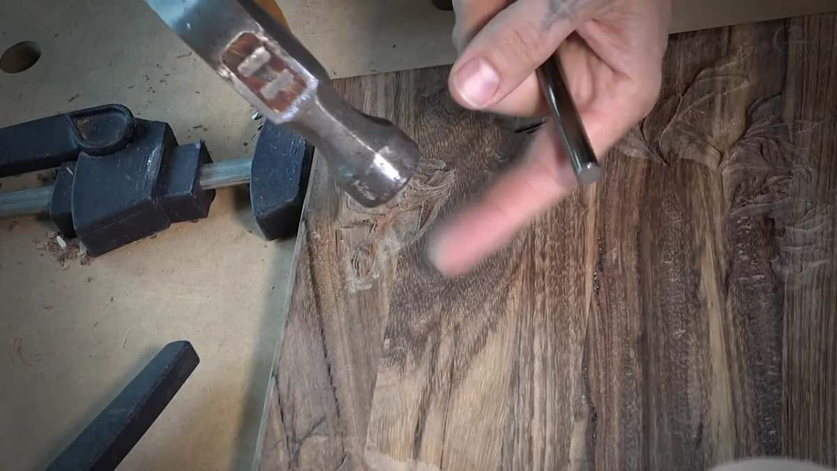 A steel hammer swings toward a punch held over dark wood, clamps along the edge