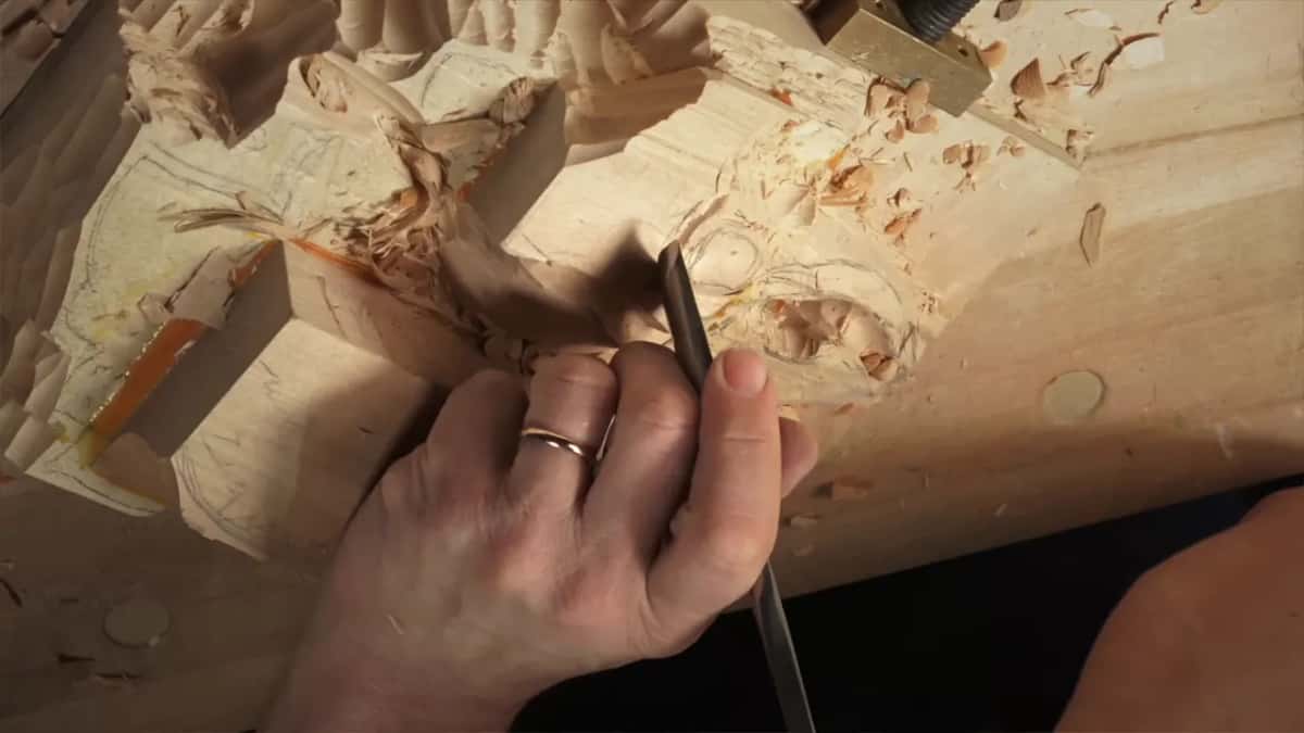 Long gouge held in both hands at the edge of a squared lump covered in pencil marks