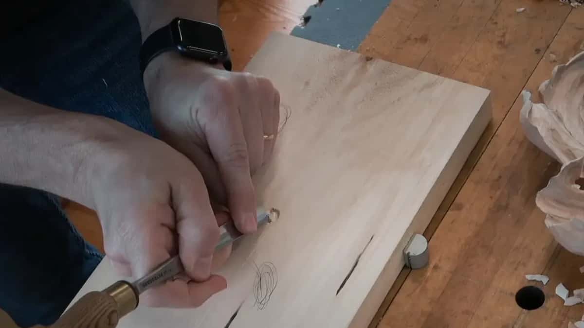 A gouge with a brass ferrule and a wood handle works at the bug, fingers guiding the blade