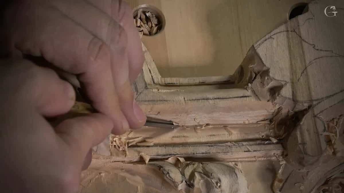 Close in, two hands push a slim tool along the bottom edge of the molding with chips around it