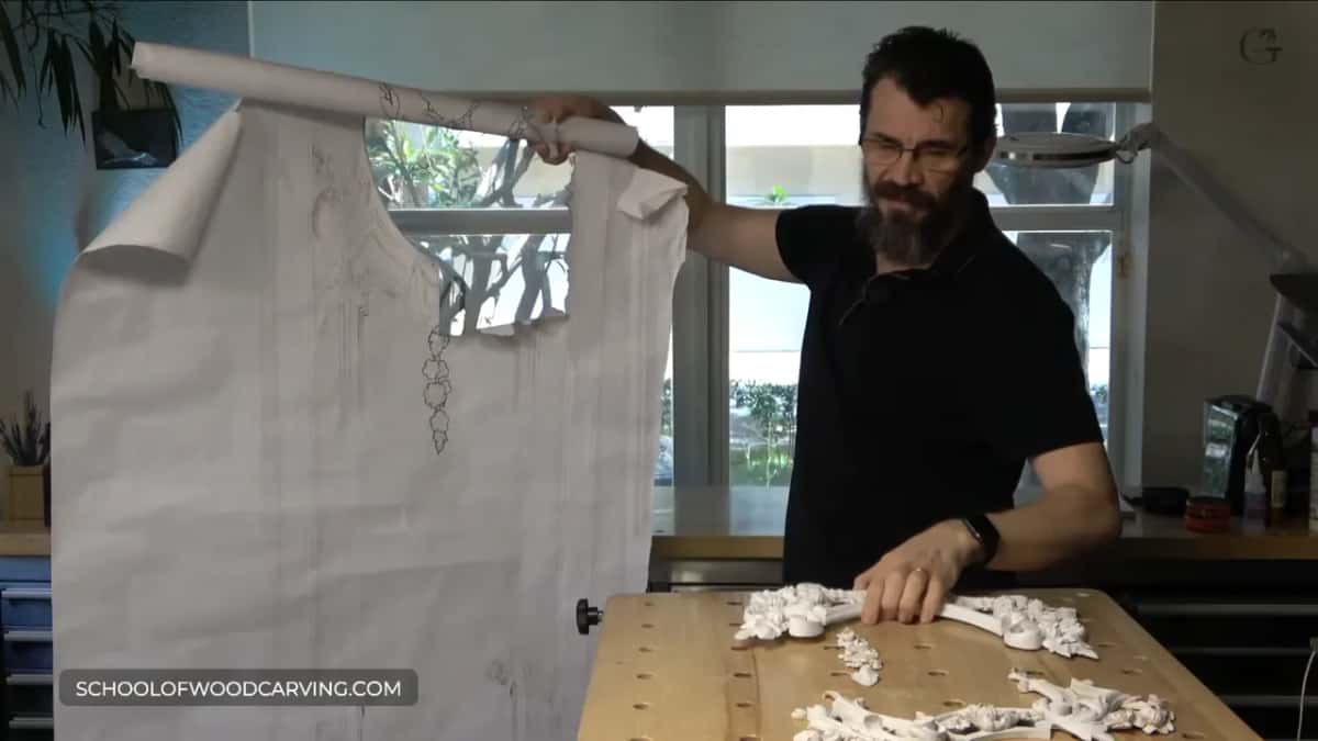 Unrolling a long paper drawing above white carvings on the bench
