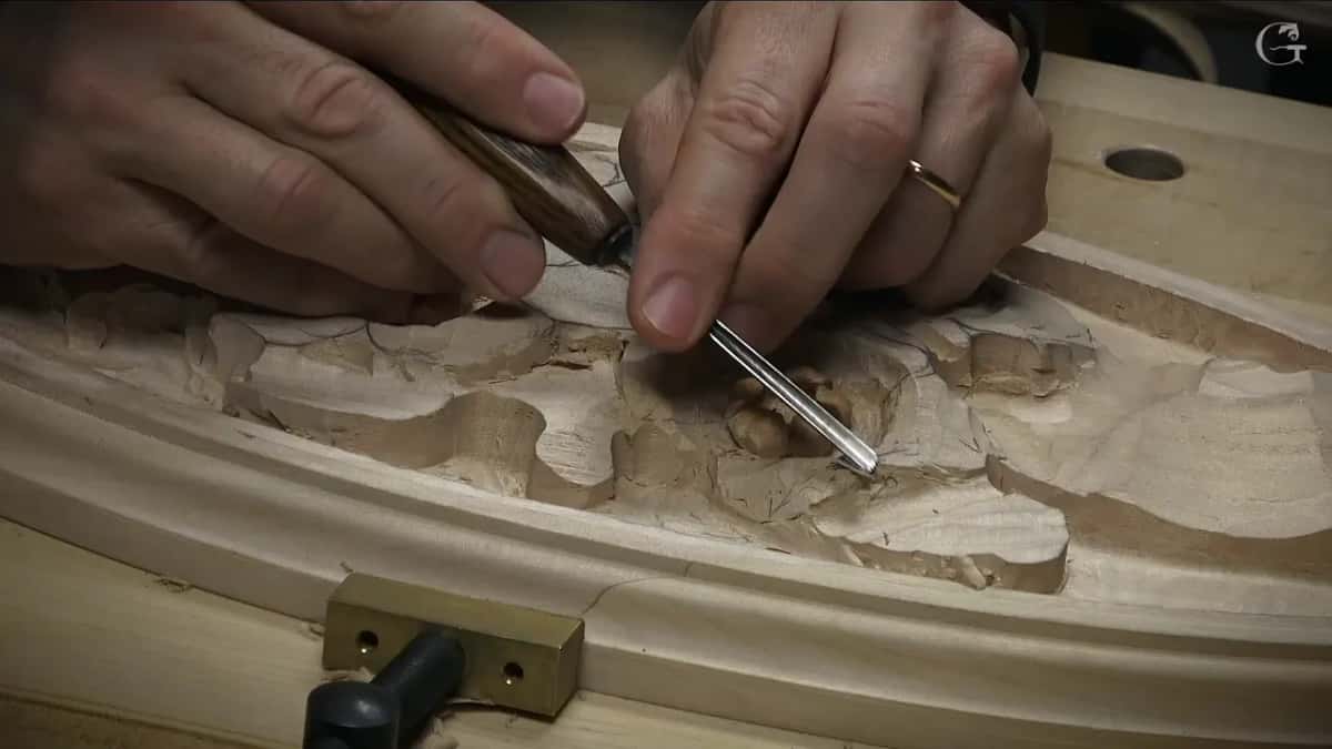 Two hands guide a slim gouge between roughed leaves near the oval rim, a brass bench dog below