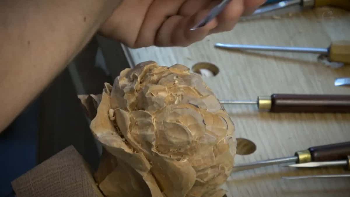 A hand holds a tool above a roughed-out flower, more tools lying on the bench beside it