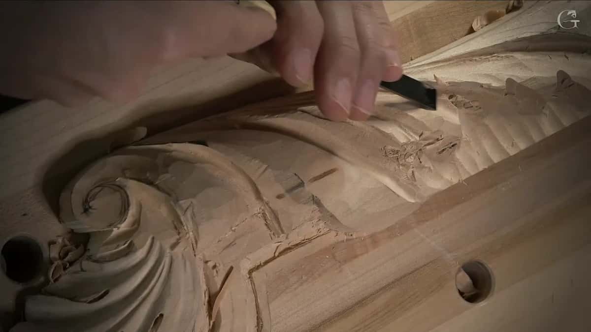 Fingers steer a narrow gouge into the leaf while the curl of the volute sits below