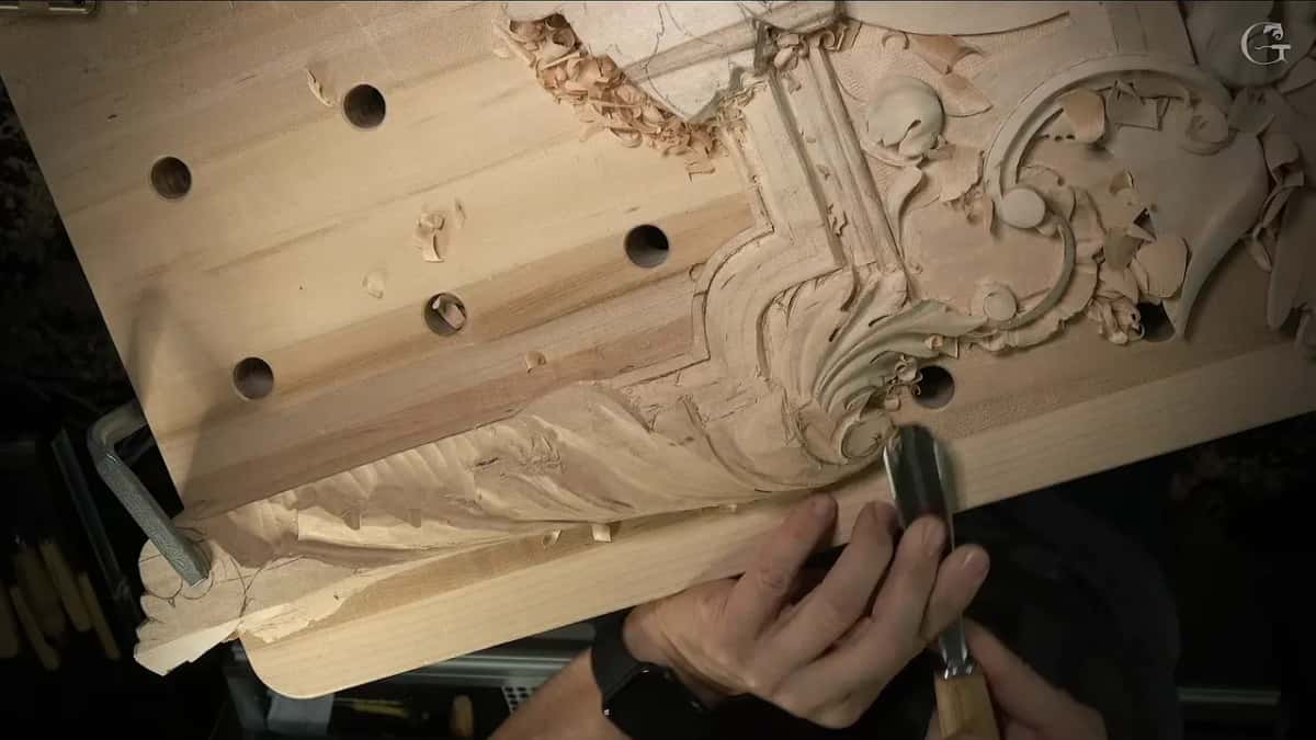 Seen from above, one hand brings a gouge down to the volute at the corner of the panel
