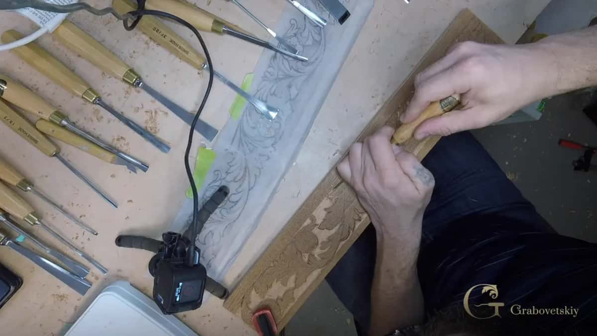 From above, both hands steer a short tool along the oak beside a row of gouges and the taped drawing