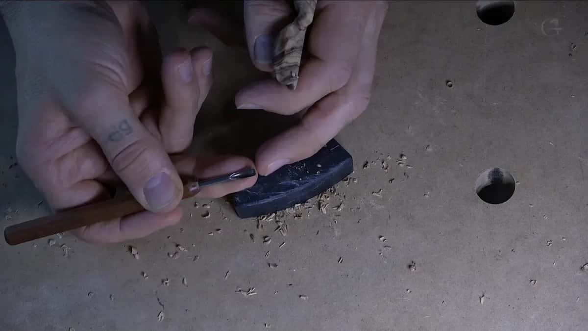 A small figured piece pinched on a dark rubber pad, a slim tool coming in near the fingers