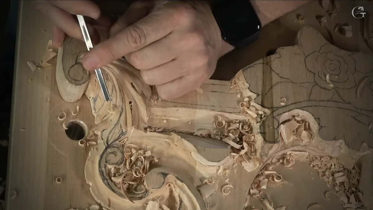 Both hands on one slim gouge, a watch on the wrist, curls of wood heaped below